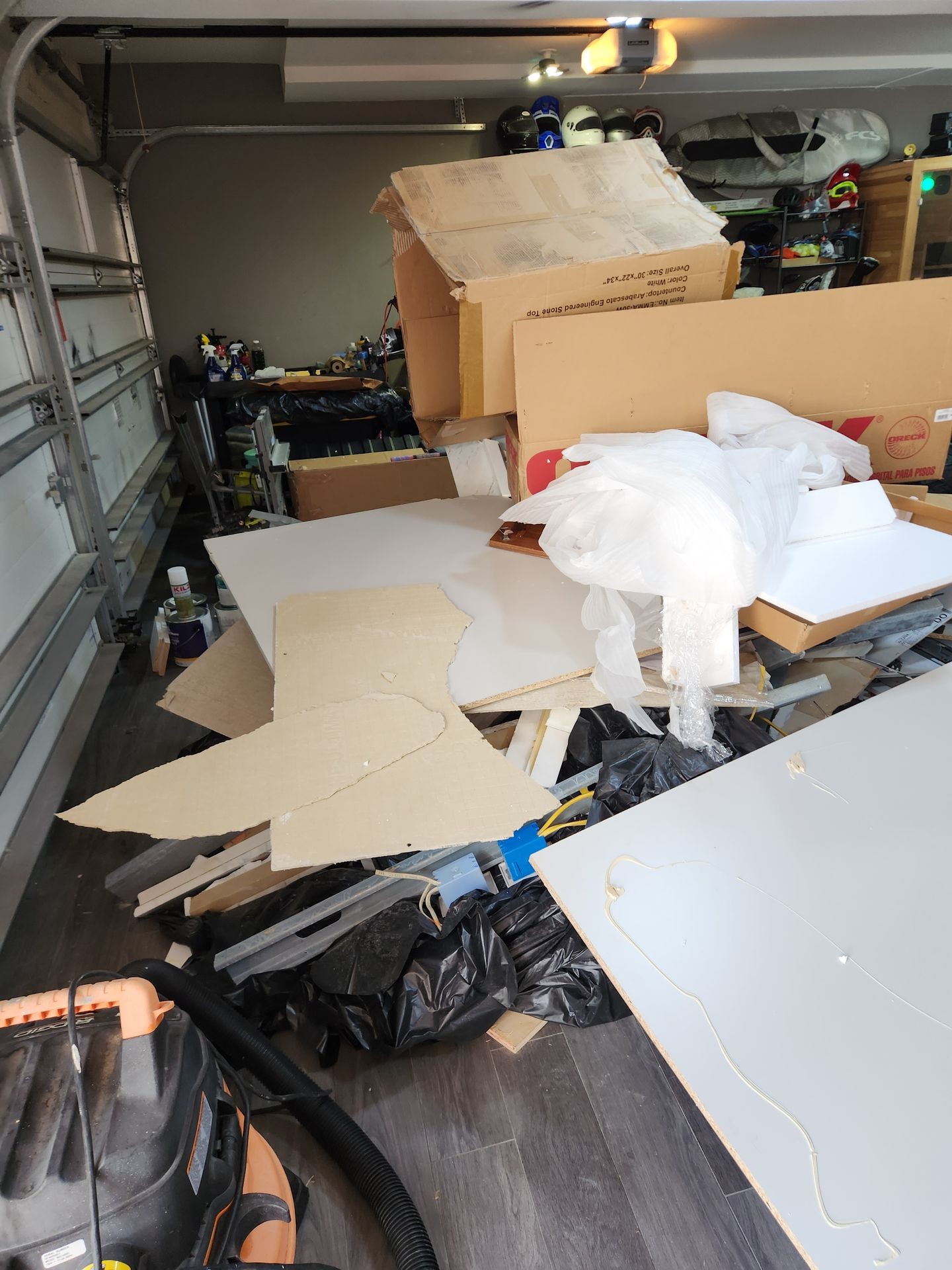 Cluttered garage with cardboard boxes, discarded packaging, and various items scattered on the floor.